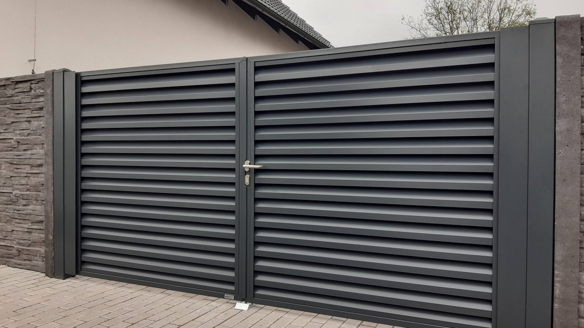 Detailní pohled na dvoukřídlou bránu (vrata) značky Nokov v antracitové barvě RAL 7016, zobrazenou z jiného úhlu, ukazující její elegantní žaluziový design